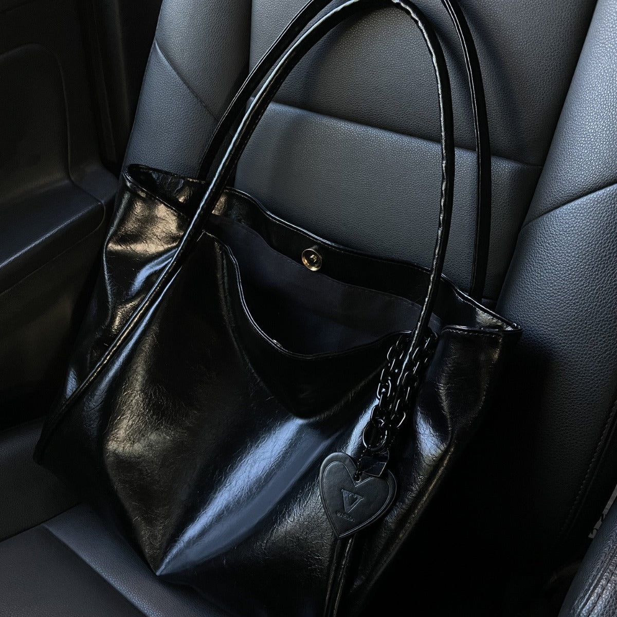 Schwarze Lacklederhandtasche mit Herzanhänger auf grauem Ledersitz | Luxuriöse Modetasche 